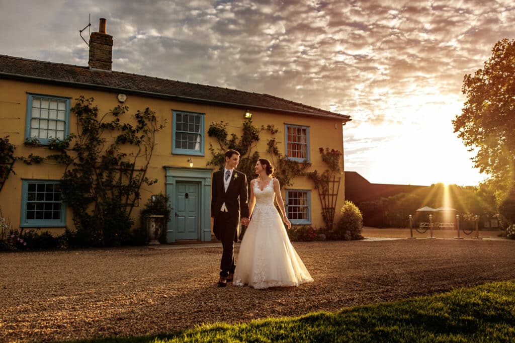 Bride and groom walk in front of country house wedding venue with beautiful sunset backdrop and pretty clouds