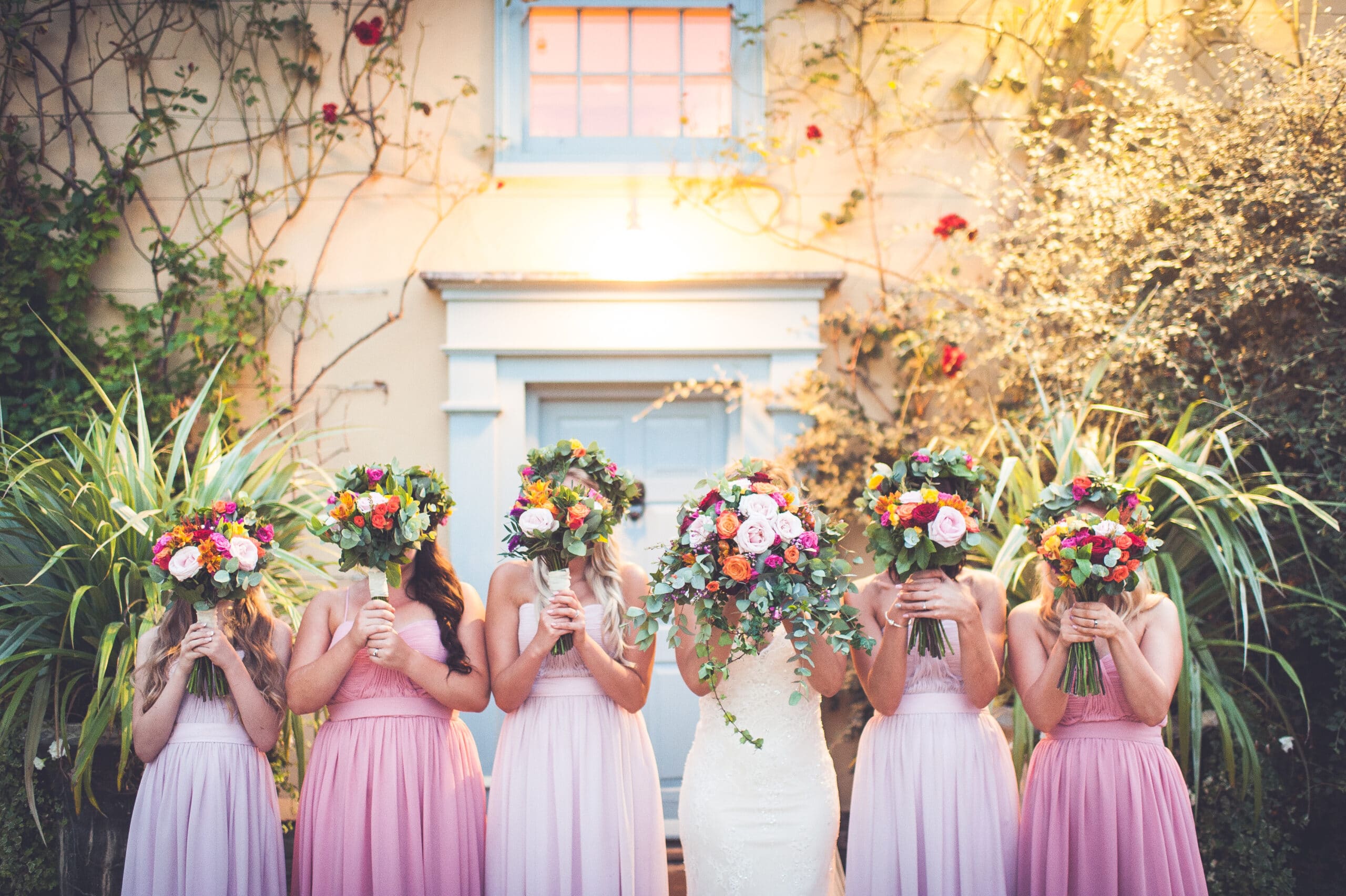 Bridesmaids and bride standing at the front of the charming Farmhouse with their bright Spring bouquets in front of their faces