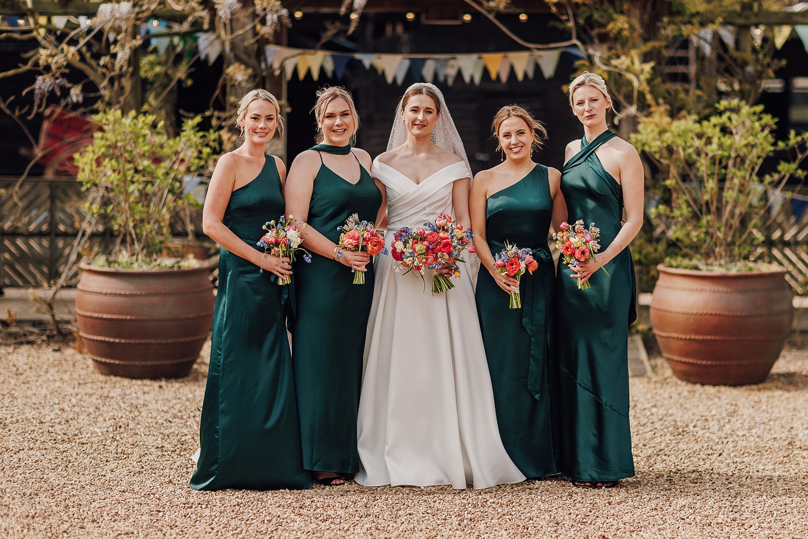 Spring wedding bride and her bridesmaids standing in the courtyard with bunting draped on the walkway leading to the rustic Tudor Barn