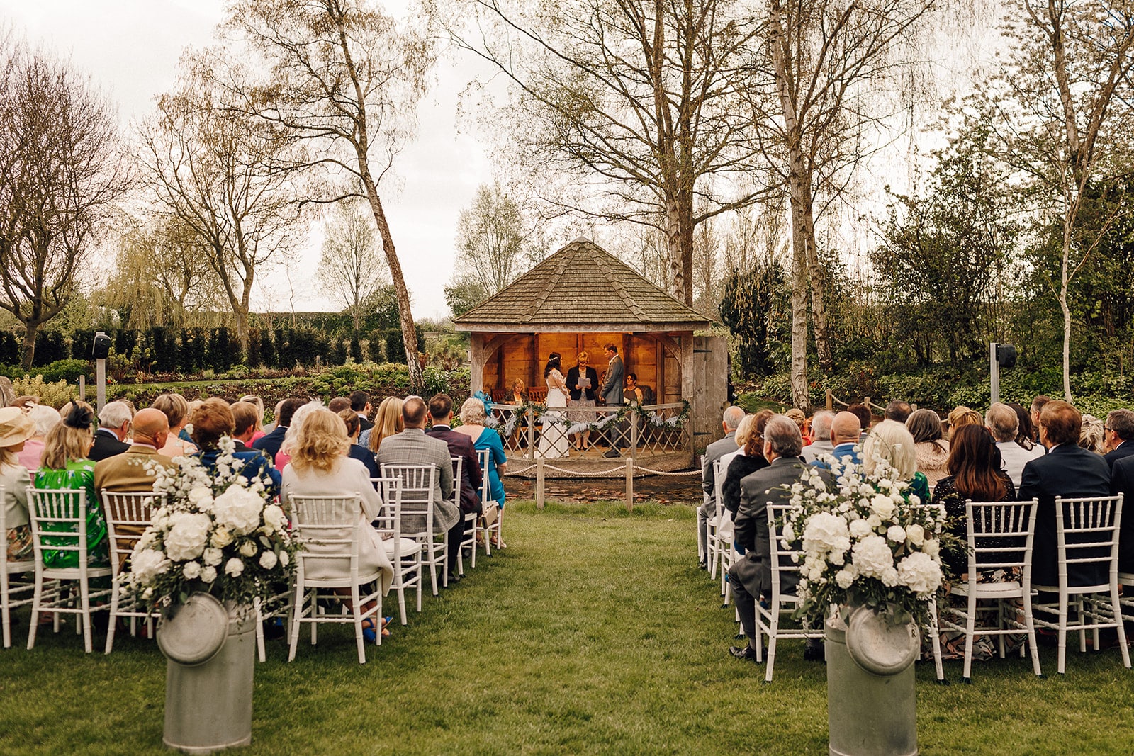 Bride and Groom saying I do in the Summerhouse with their guests watching from the pond banks with Spring garden in the background