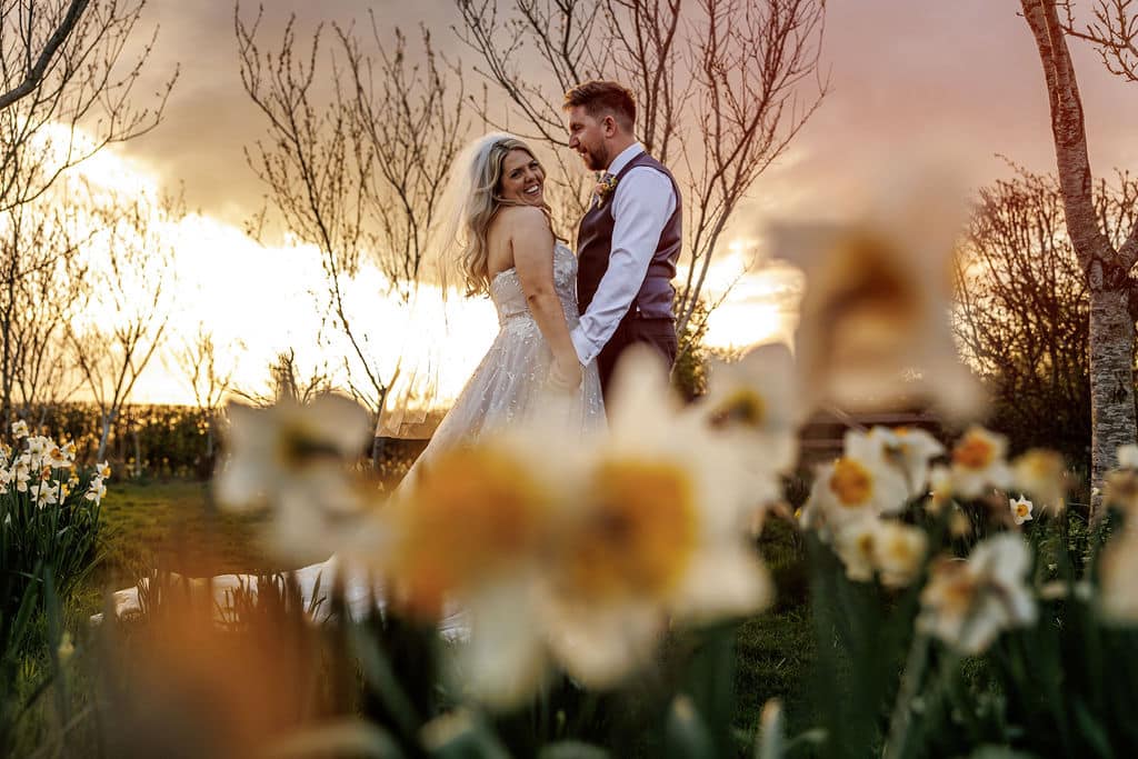 Spring wedding garden at outdoor wedding venue as bride and groom stand in field of daffodils surrounded by trees