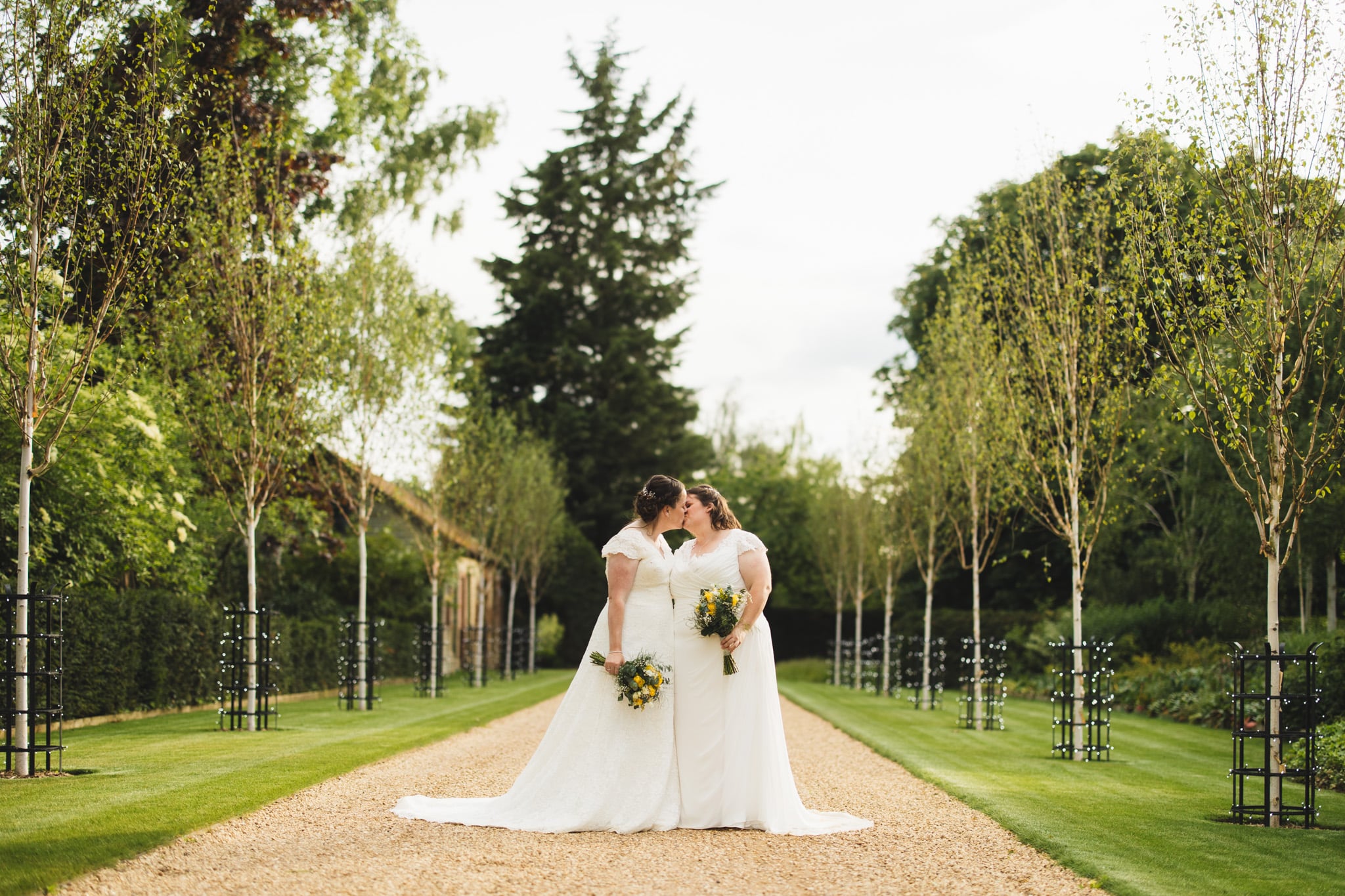 Two brides same sex wedding share kiss at tree lined driveway of countryside wedding venue