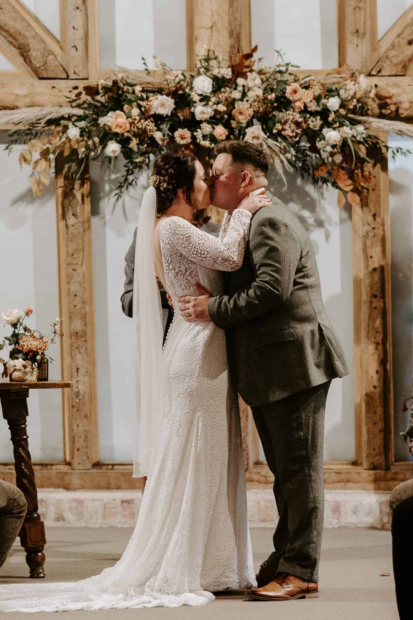 Couple kiss after just marrying in barn wedding ceremony with beautiful autumn flower garland on rustic wooden beams