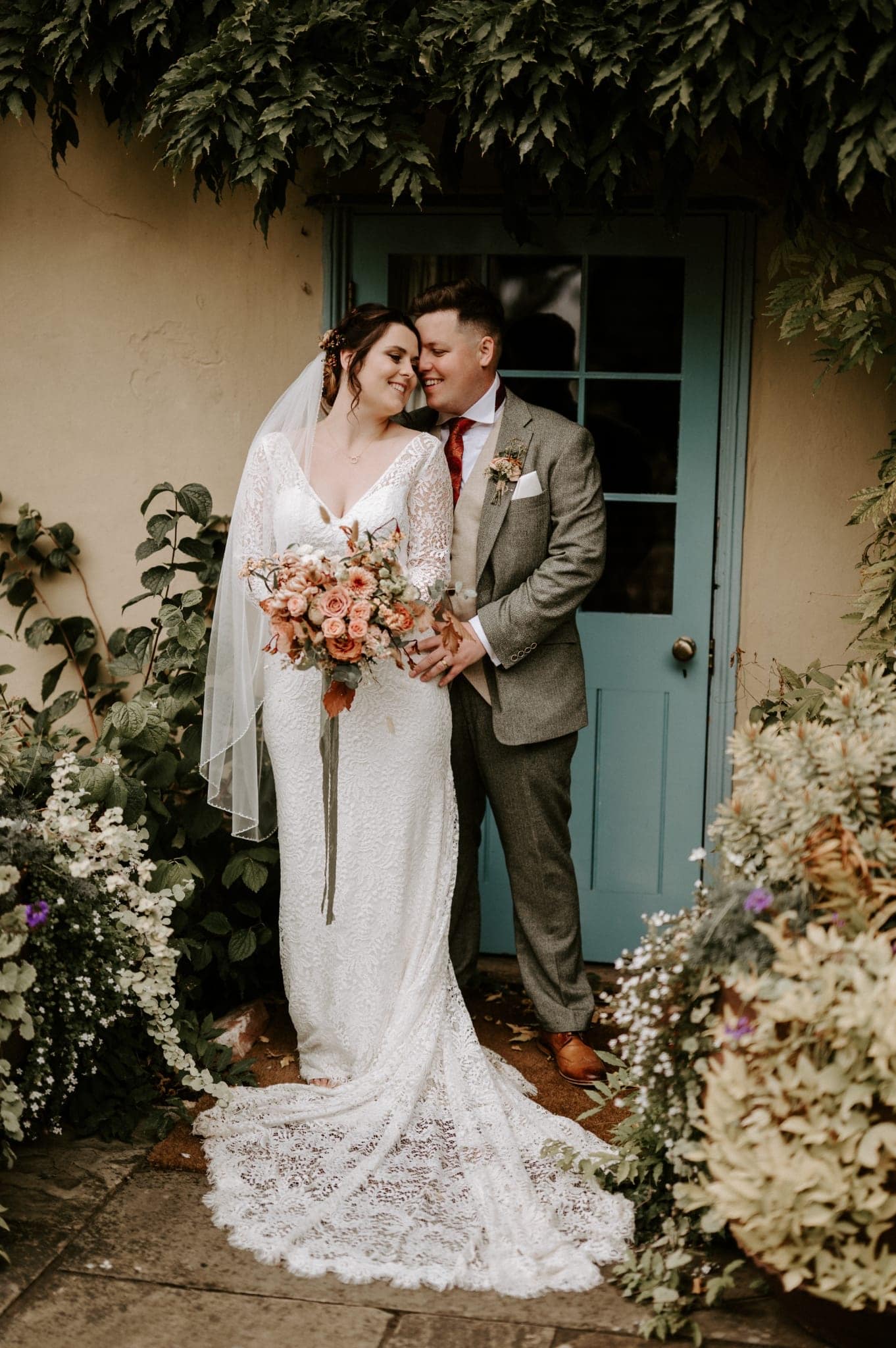 Bride in wedding gown and groom in tweed suit kiss in front of blue farmhouse door