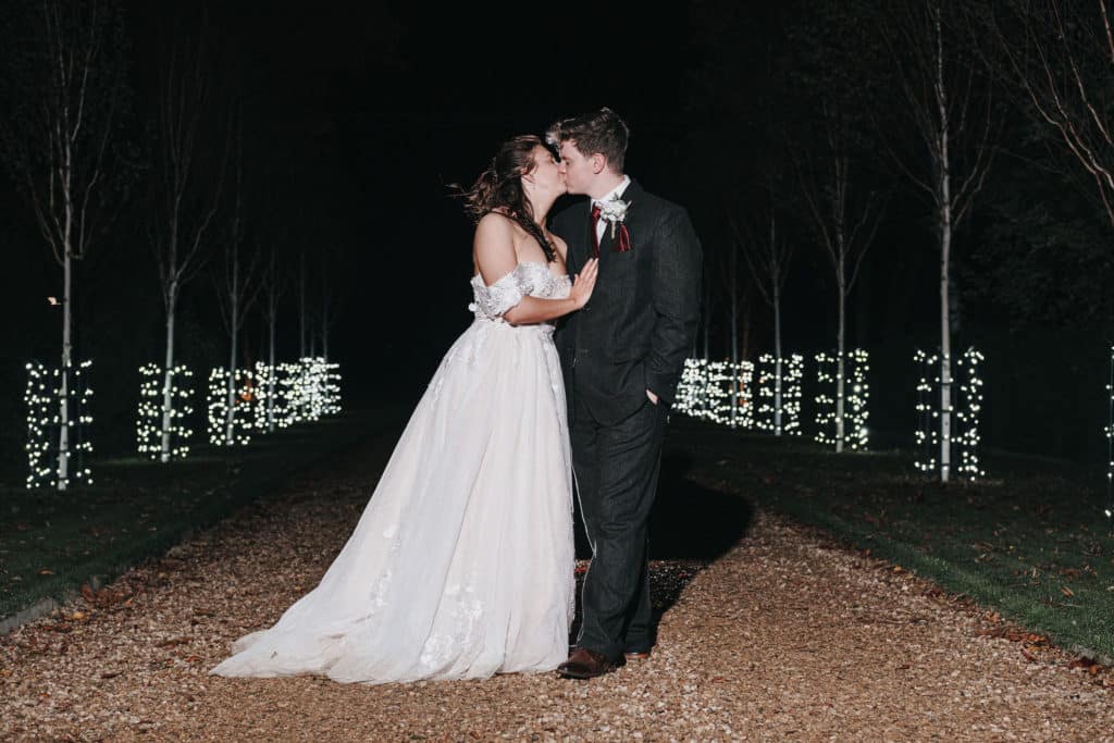 Bride and groom share a kiss at countryside wedding venue on tree lined driveway lit by fairy lights