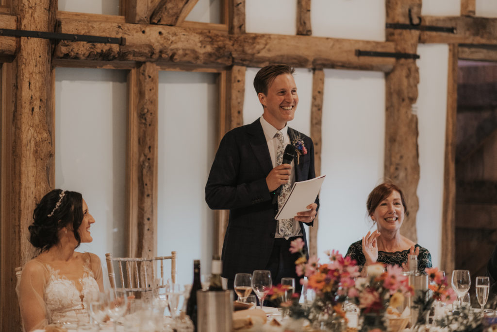 Groom makes a speech at barn wedding venue as bride and guests look on