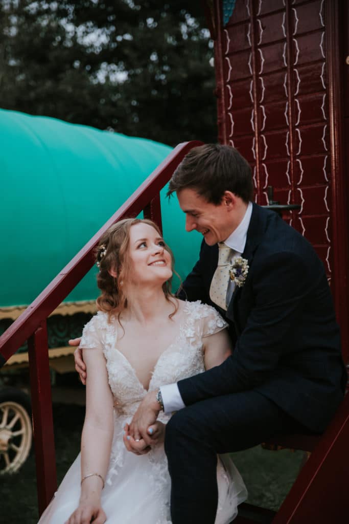 Bride and groom just married share a moment on steps of colourful romany glamping wagons at countryside wedding venue 