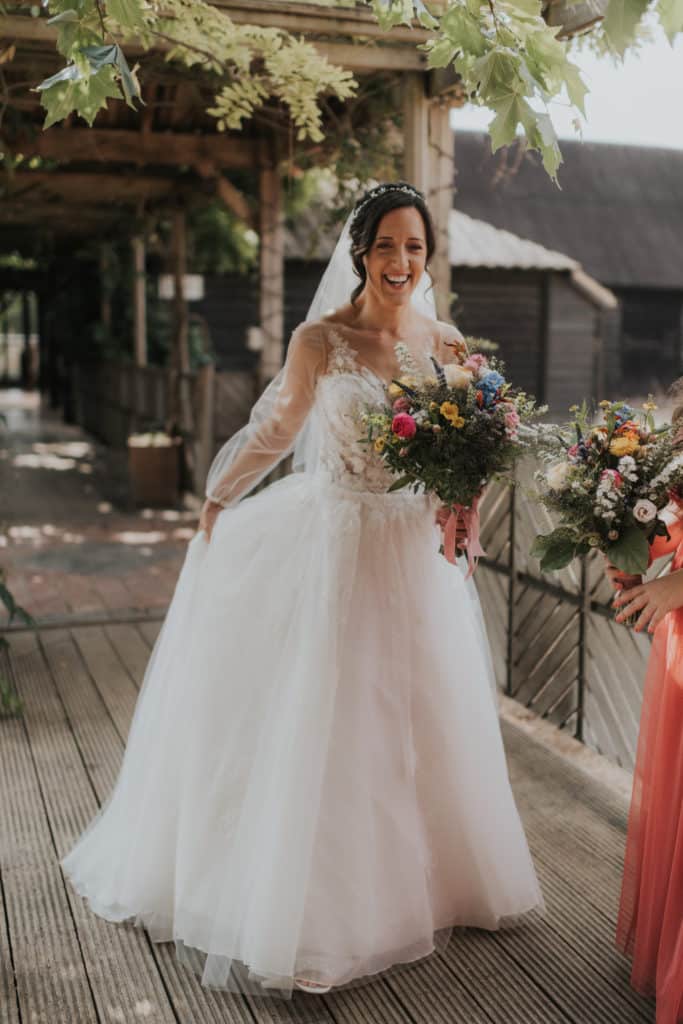 Stunning bride in beautiful wedding dress carries bouquet on wooden walkway of countryside wedding venue