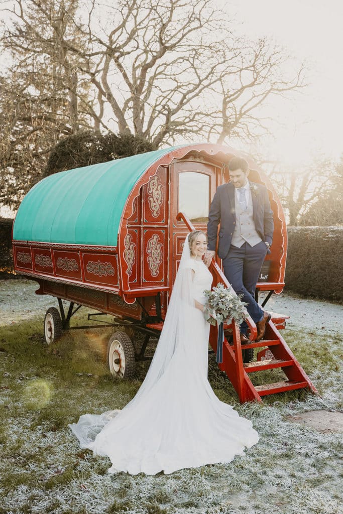 Couple at quirky winter wedding venue on frosty day in front of Red Romany Wagon at countryside wedding venue 