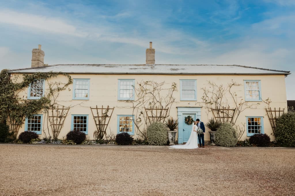 Bride and Groom on Wedding day in front of cream country farmhouse with blue door and windows and christmas wreath on door