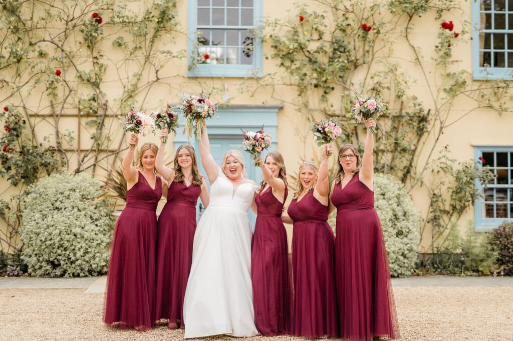 Bride in beautiful white wedding dress and bridesmaids in burgundy dresses celebrate on wedding day in front of pretty county farmhouse