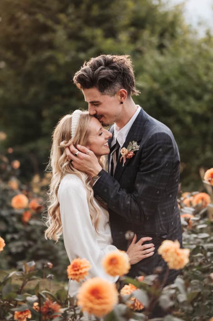 Groom kisses bride in wedding day with colourful peach flowers in foreground