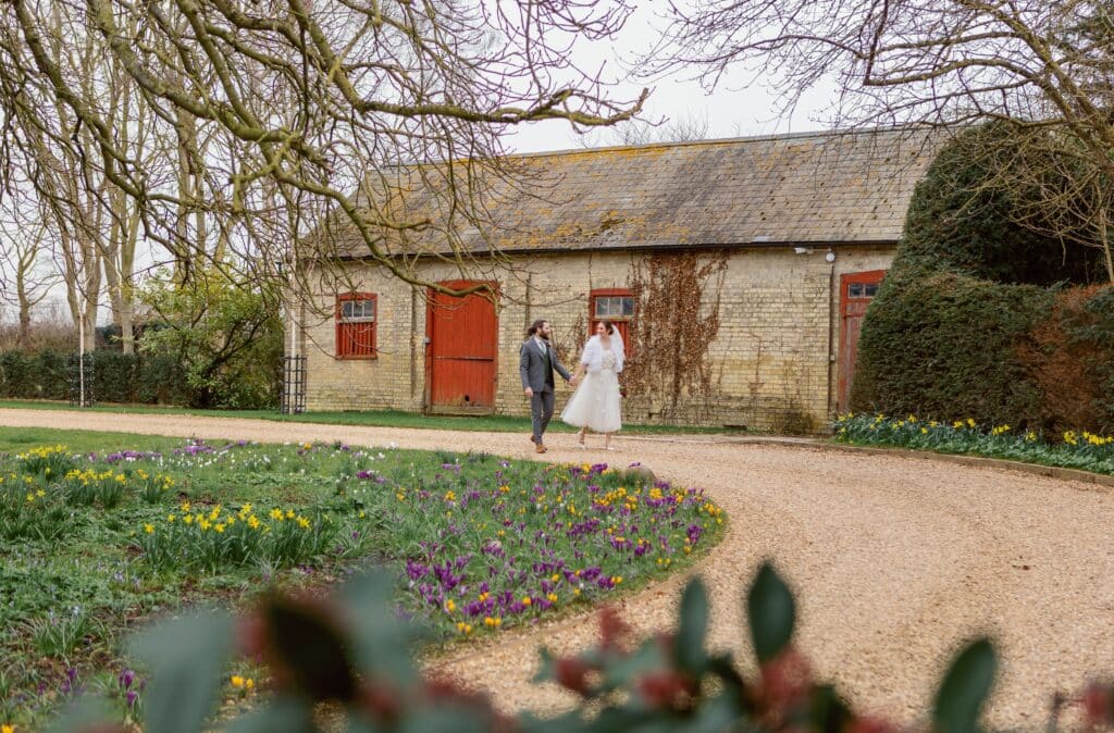 Bride and Groom at Outdoor Spring Wedding venue walking past red barn doors and colourful spring flowers 