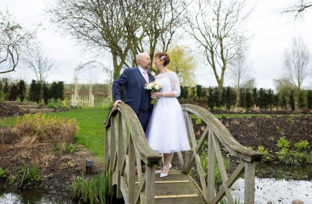 Photo of bride in white dress and groom in blue suit on bride over pond at garden wedding venue
