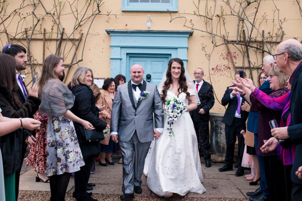 Just married bride and groom enjoy a confetti shower with their guests in front of cream farmhouse wedding venue with blue door 
