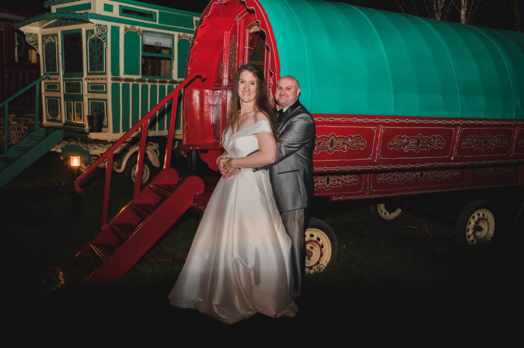 Bride and Groom in front of colourful red and green romany wagon at Jewish wedding venue in the countryside