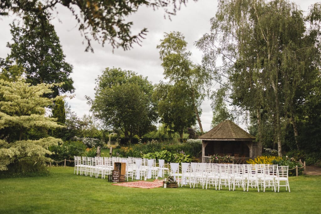 Garden wedding venue set for ceremony with white ceremony chairs a centre aisle with antique rugs and a pretty summerhouse adorned with flowers 