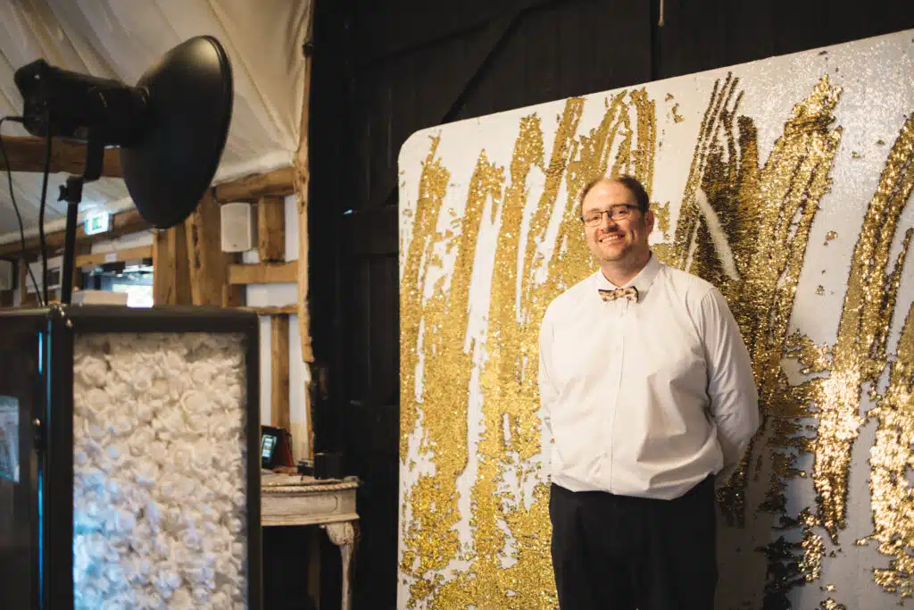 Owner at Photo Booth in front of booth and gold backdrop 