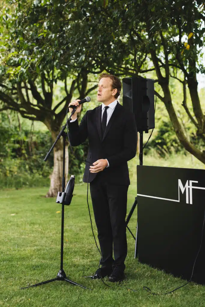 Male swing singer performing in black suit and tie in garden of wedding venue 