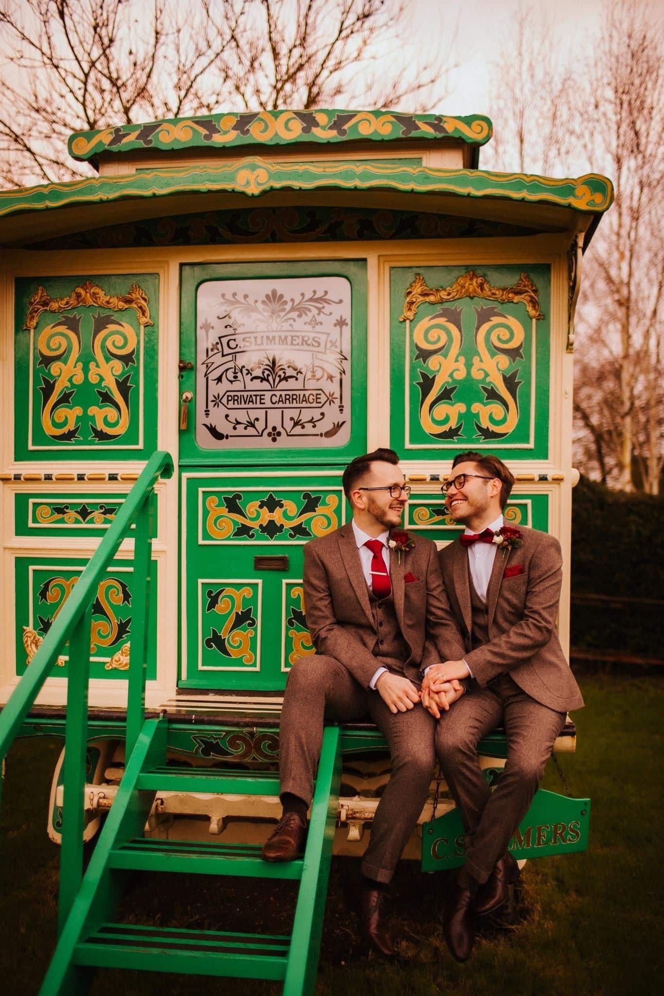 Same sex wedding two grooms in tweed suits on wedding day sitting on steps of cream and green showman's wagon