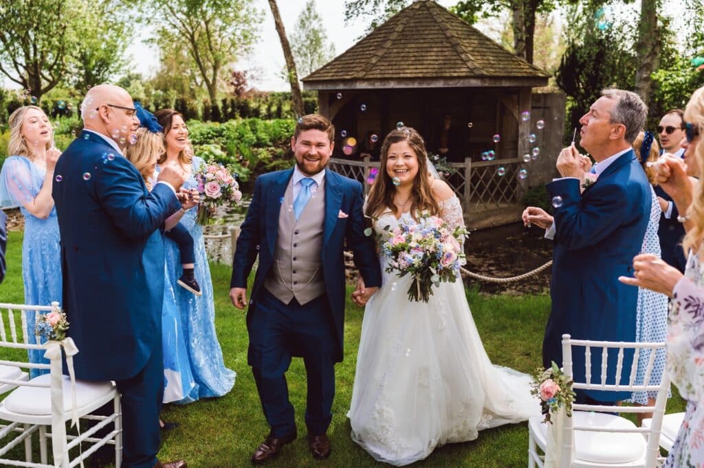 Bride and Groom exit aisle after garden wedding ceremony surrounded by their guests blowing bubbles for the photo