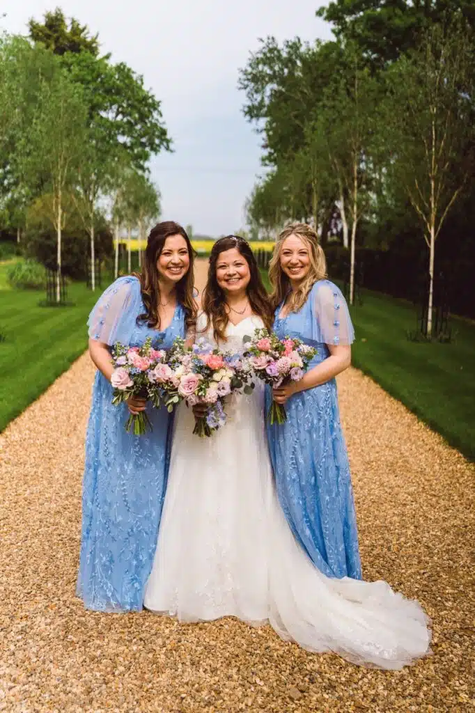 Bride in white wedding dress and two bridesmaids in beautiful blue dresses smile for photo on treelined driveway