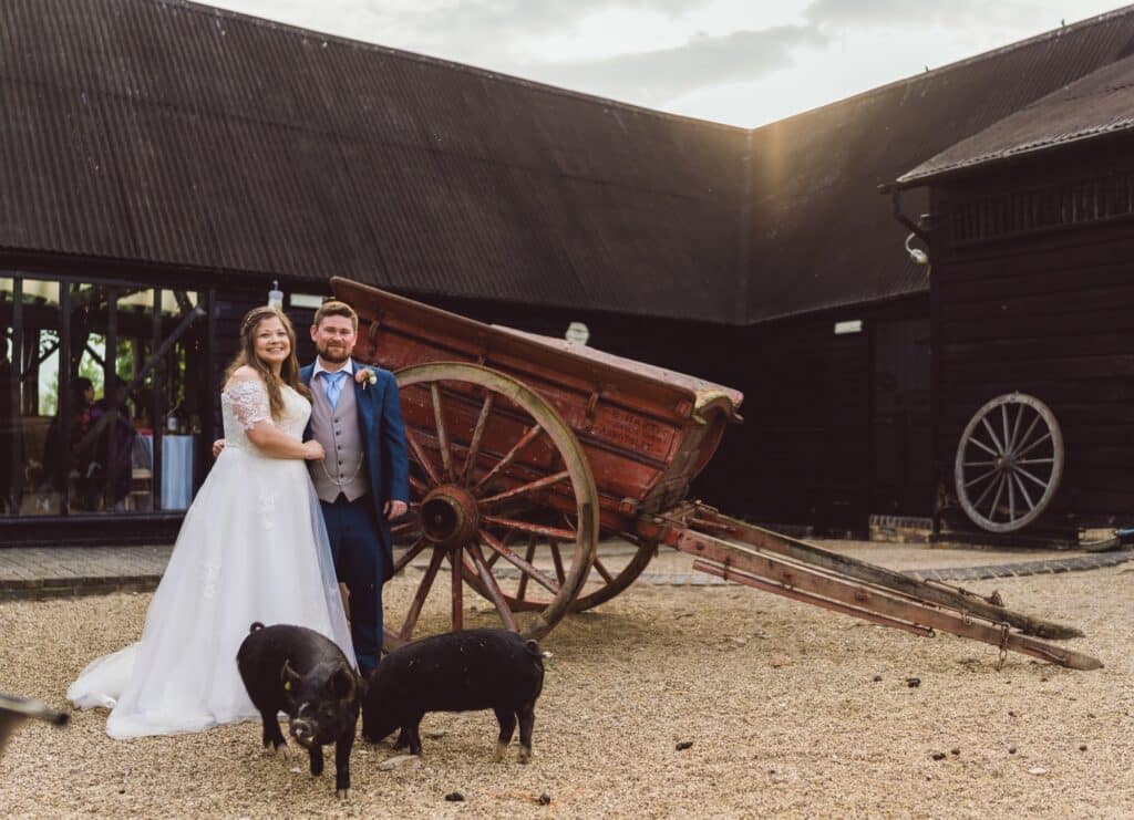 Bride and groom pose for wedding photo in front of farm carriage with piglets in the foreground at farm wedding venue