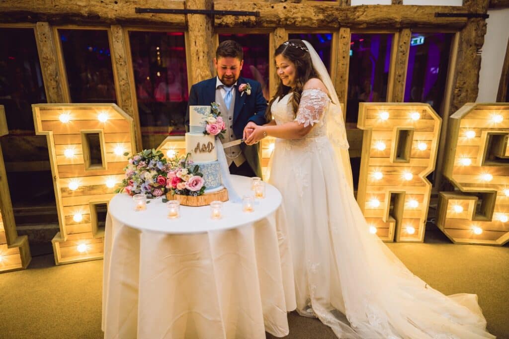 Bride and groom cut wedding cake in barn wedding venue in front of mr & mrs light up letters 