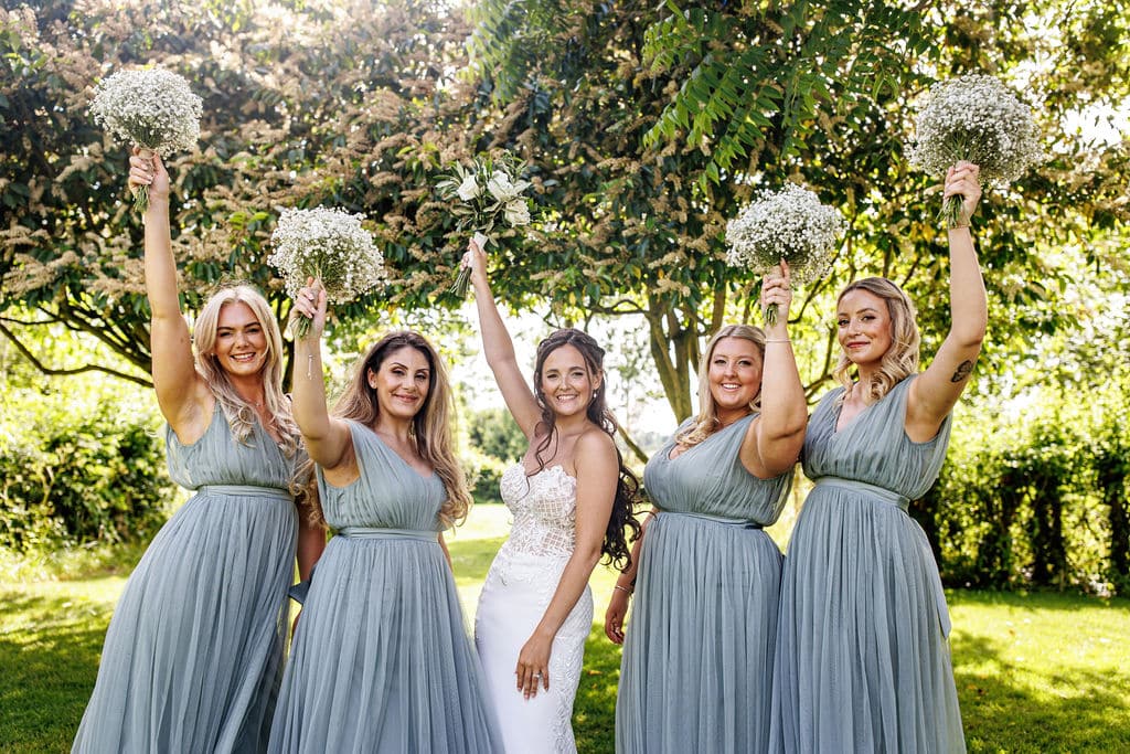 Bride and bridesmaids in garden wedding venue raise bouquets in celebration