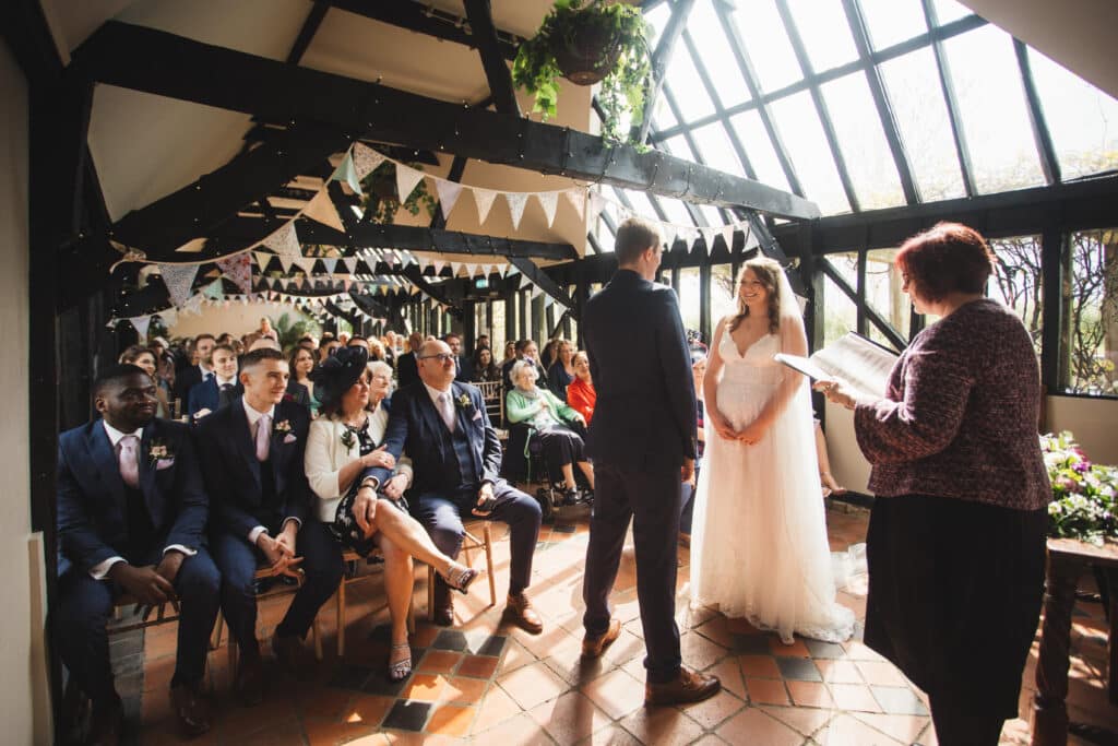 Bride and Groom say their wedding vows at barn ceremony with black timber beams and sunlit sky lights  in front of guests 