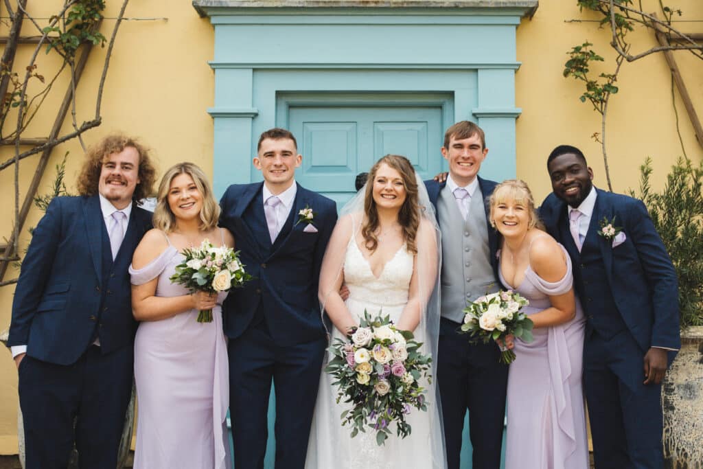 Bride and groom with their groomsmen and bridesmaids in front of cream farmhouse with blue door 