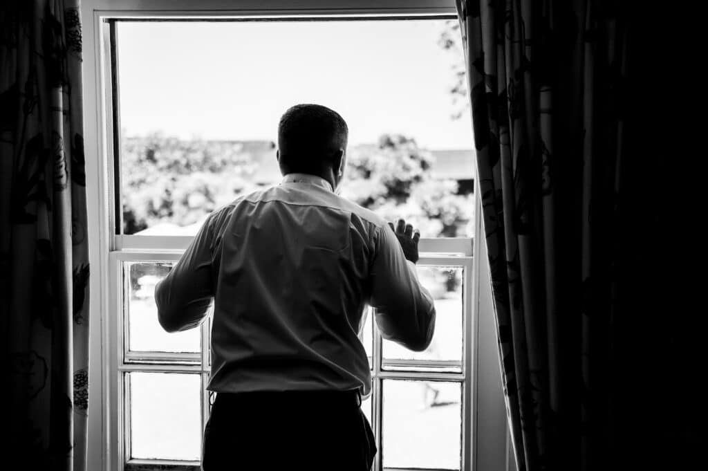 Bridegroom looks out of window at countryside wedding venue on morning of wedding day black and white image 