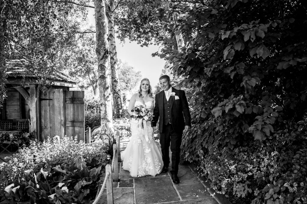 Black and white photo of Just married bride and groom at outdoor ceremony walking hand in hand under trees 