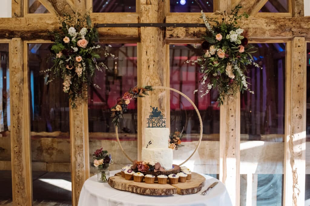 Wedding cake at barn wedding venue with flowers on beams and floral hoop behind wedding cake