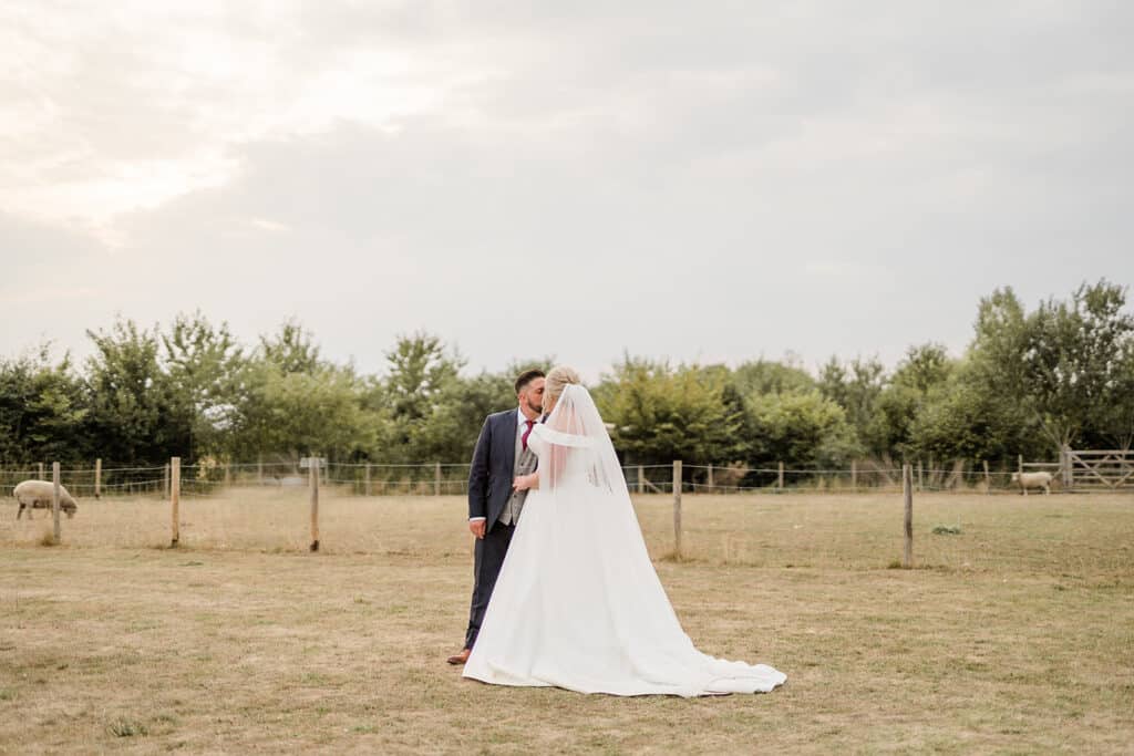 Farm Wedding Venue Bride and groom standing in meadow on sunny day with sheep in the background