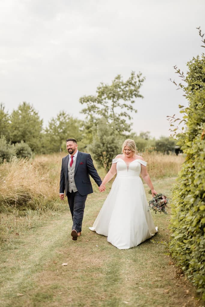 Bride and groom on wedding day at countryside wedding venue walking hand in hand past hedgerow