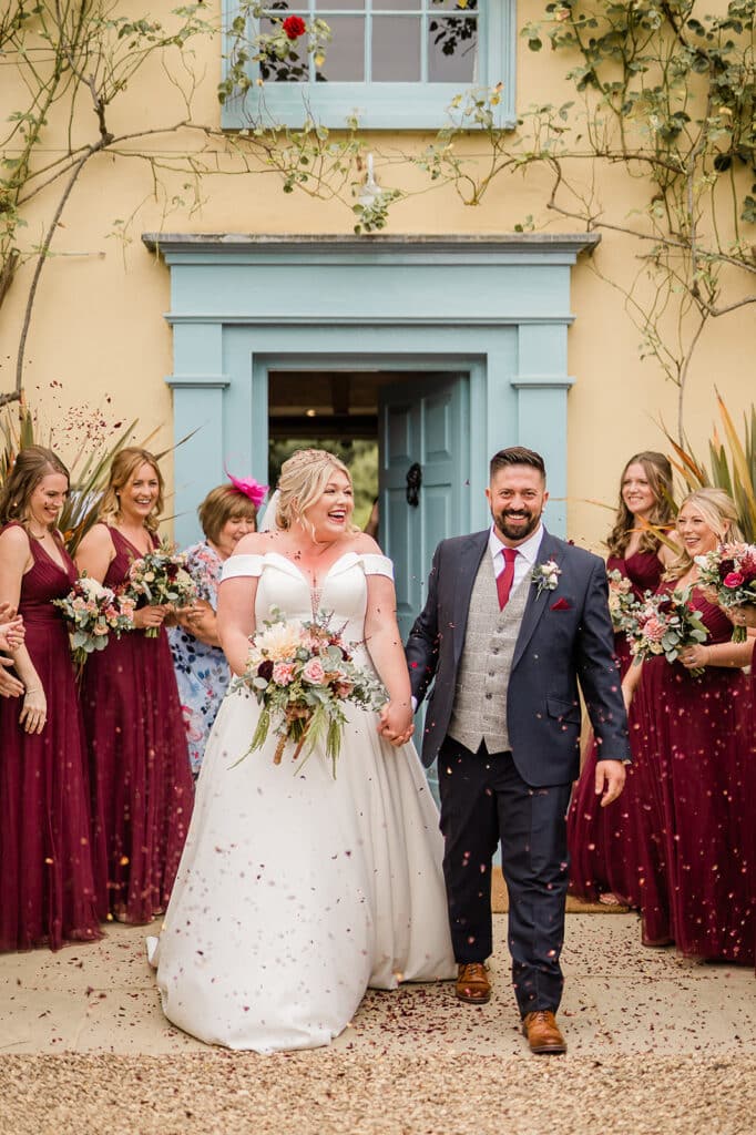 Bride and groom in front of pretty cream farmhouse with blue open door walking past bridesmaids in deep red dresses