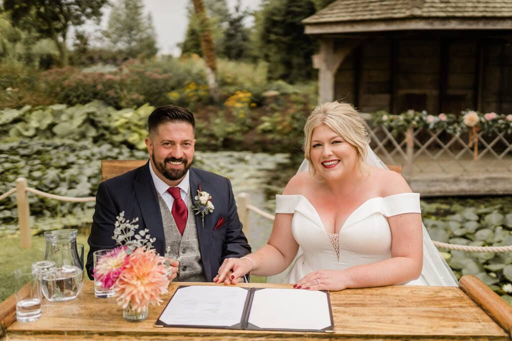 Bride and groom smile for photo as they sign marriage register with summerhouse and pond in the background