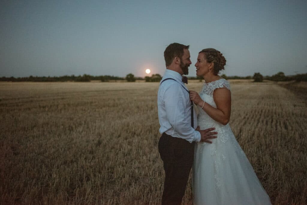 Bride and groom at farm wedding venue in field of corn as sun goes down behind them 