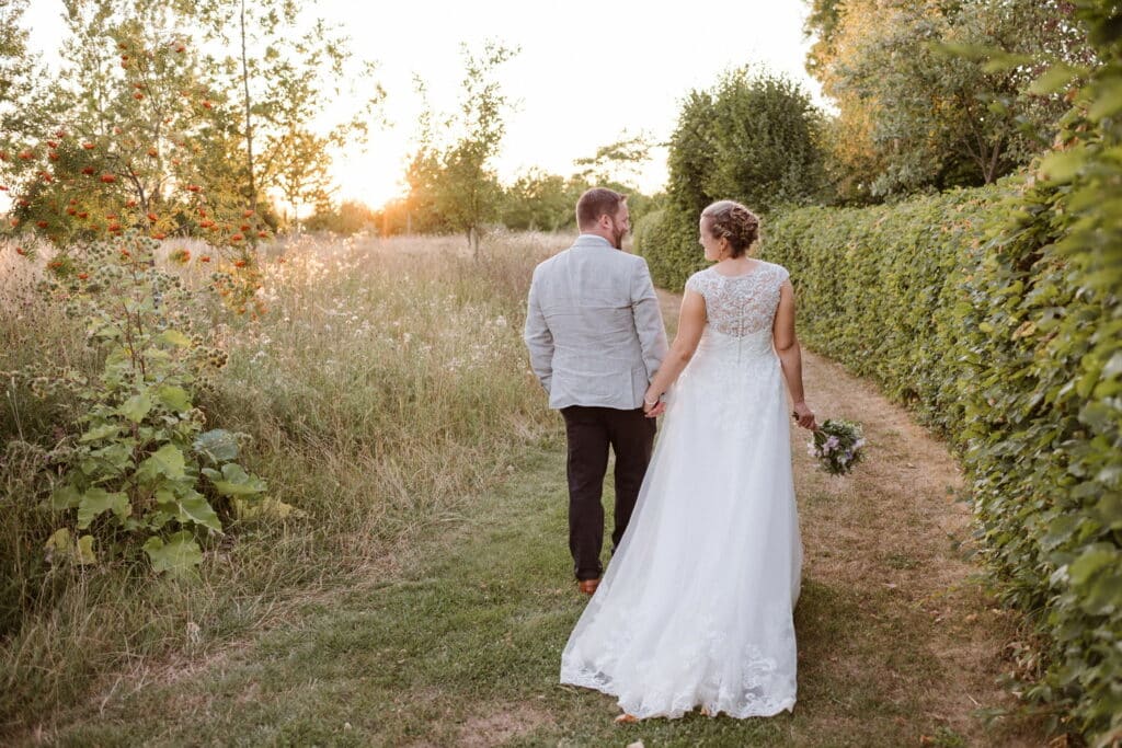 Bride and groom walk hand in hand away from the camera in garden wedding venue at sunset 