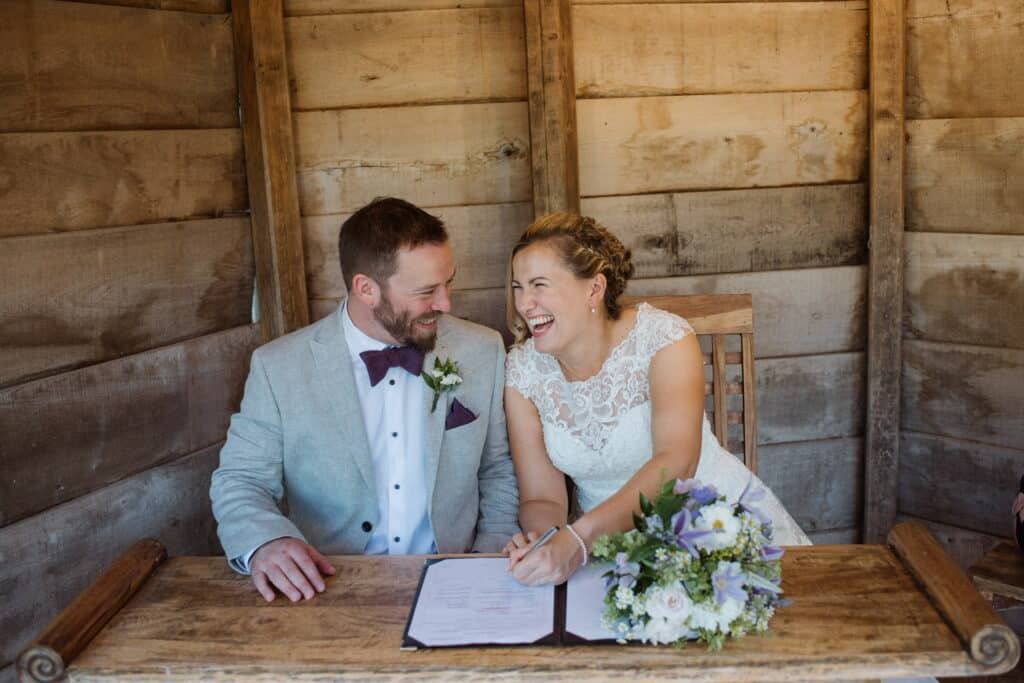 Bride and groom smiling and laughing as they sign their wedding certificate in pretty wooden summerhouse