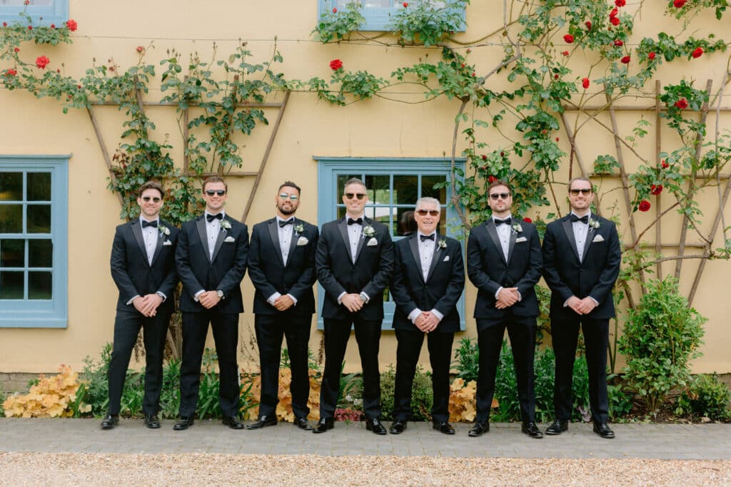 The groom and his groomsmen standing at the front of country farmhouse adorned with bright red roses