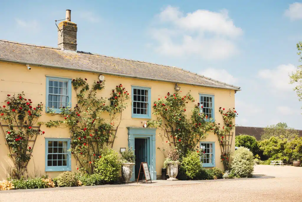 Photo of countryside wedding venue on sunny day cream and blue farmhouse with red roses growing by door