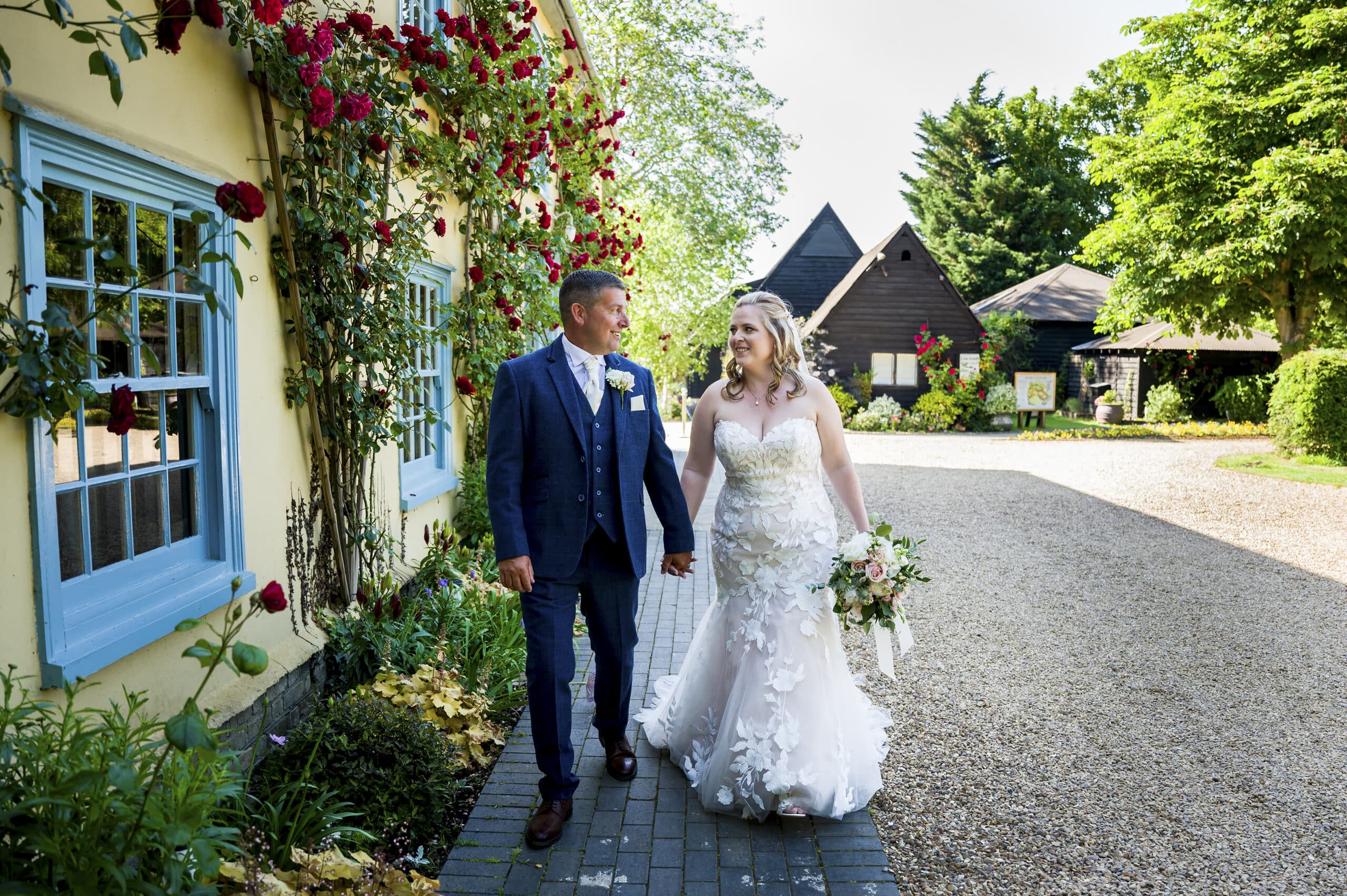 Bride and groom on sunny wedding day hold hands and walk past pretty country house wedding venue