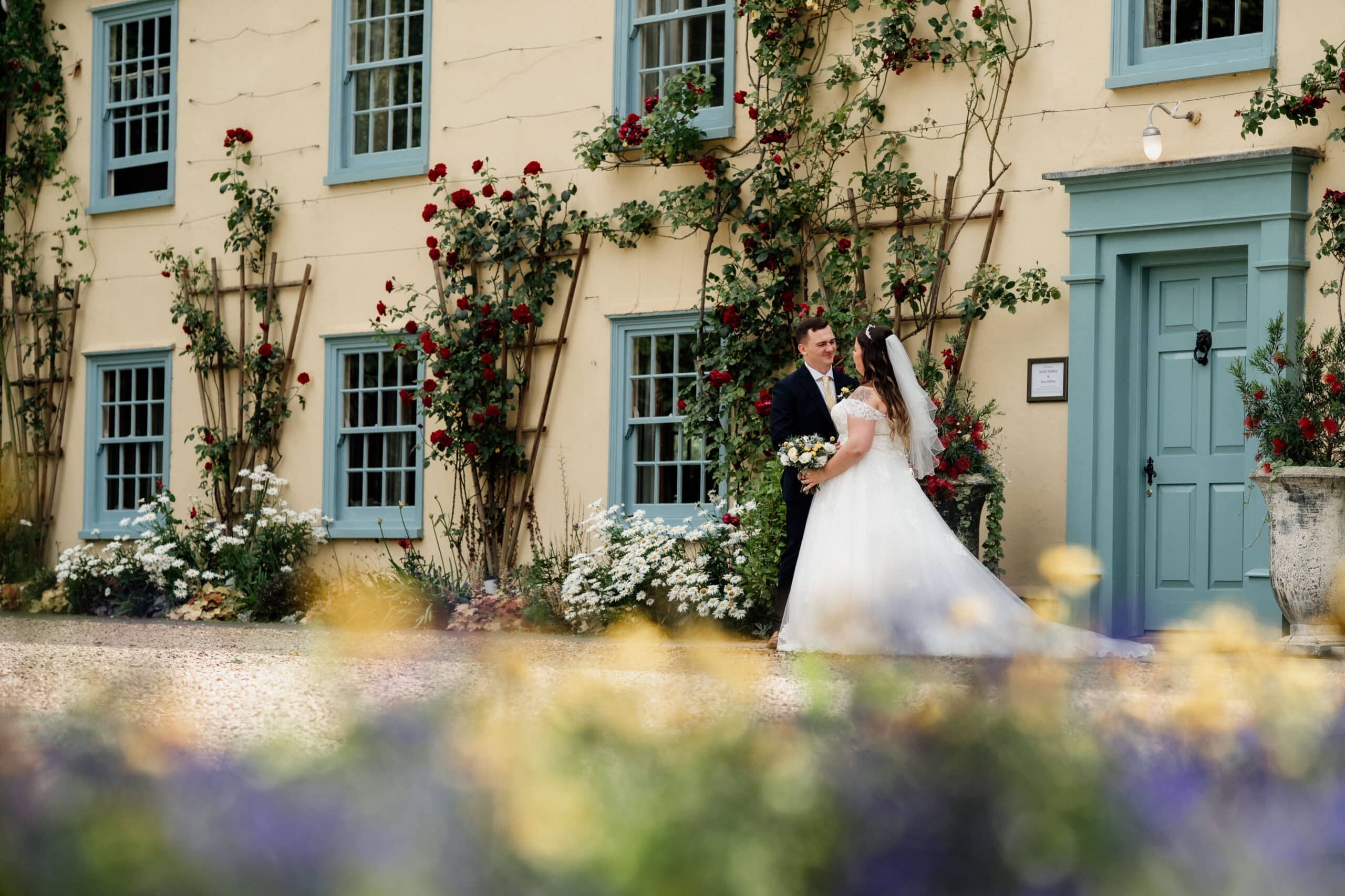 Bride and groom in front of cream country farmhouse with blue windows and red roses round the door