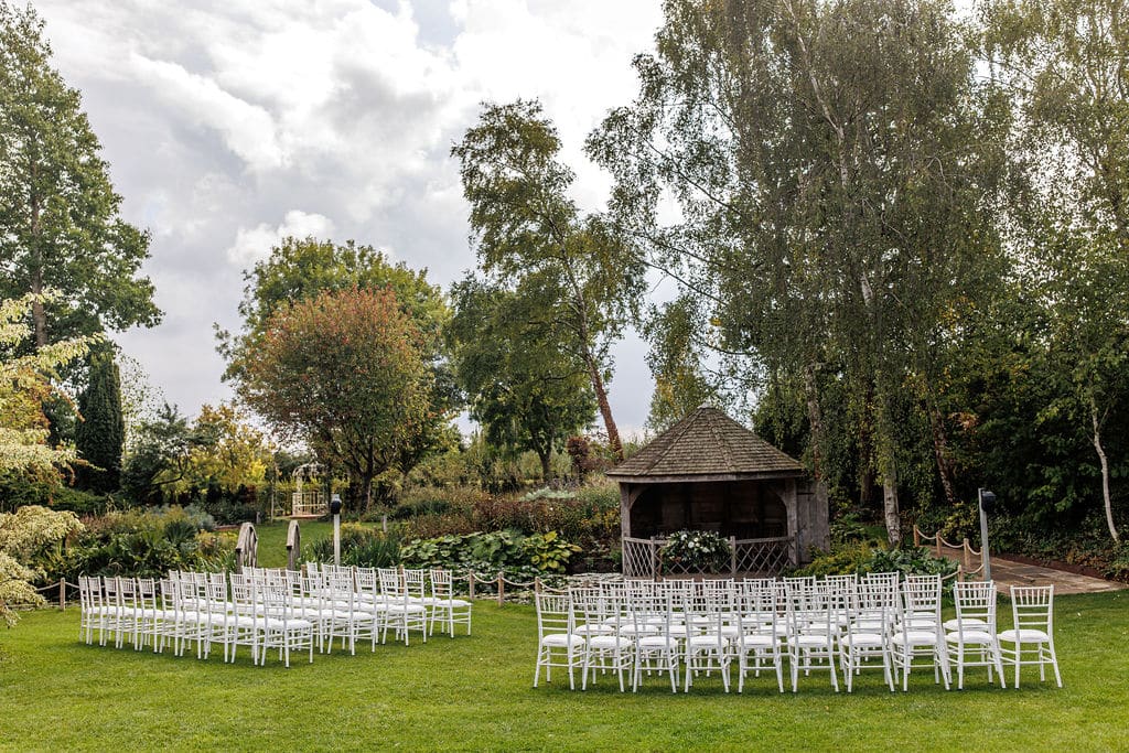 Garden wedding ceremony with white ceremony chairs in front of pretty rustic wooden summerhouse and tall trees