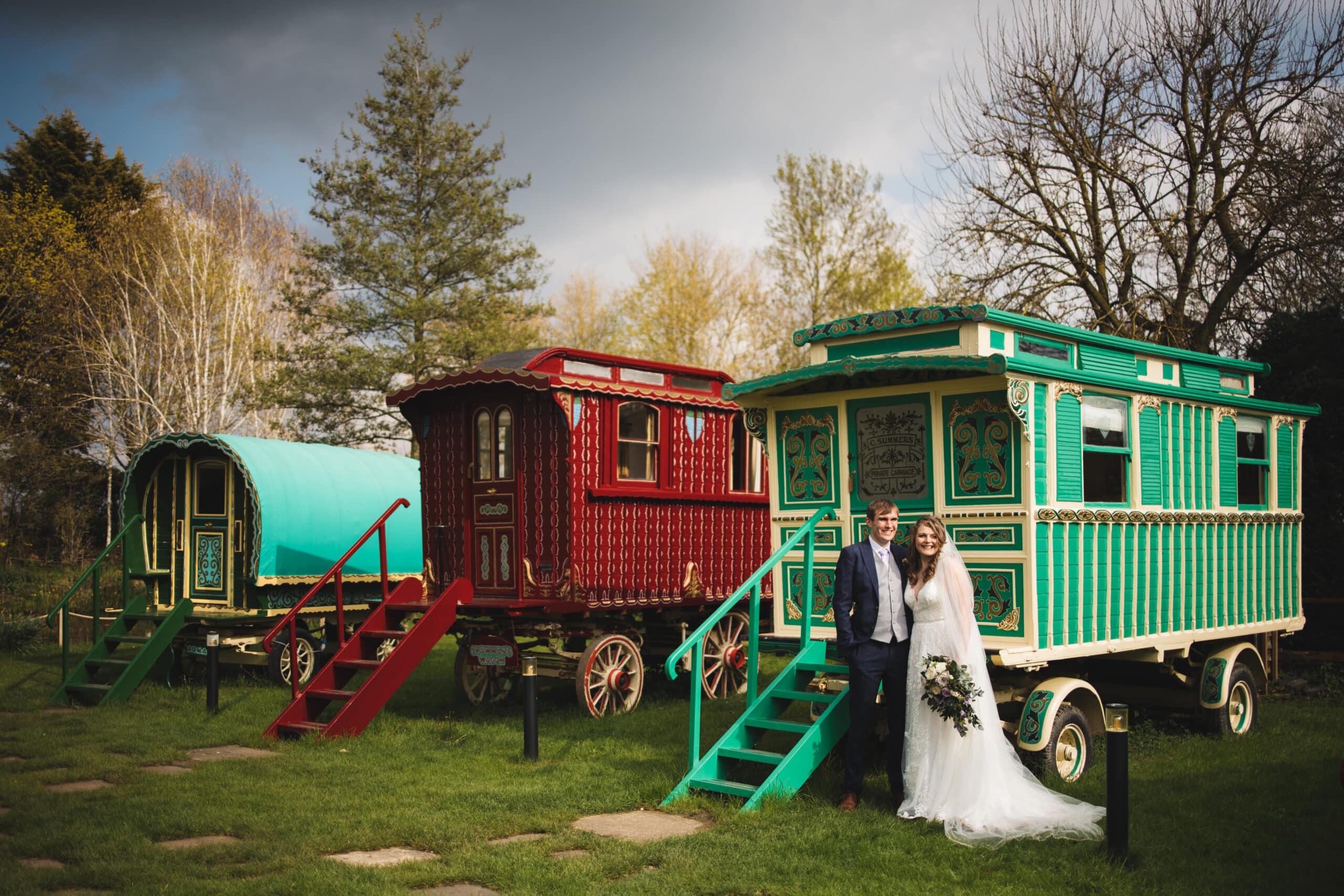 Bride and Groom in front of colourful green and red romany wagons at rustic countryside wedding venue