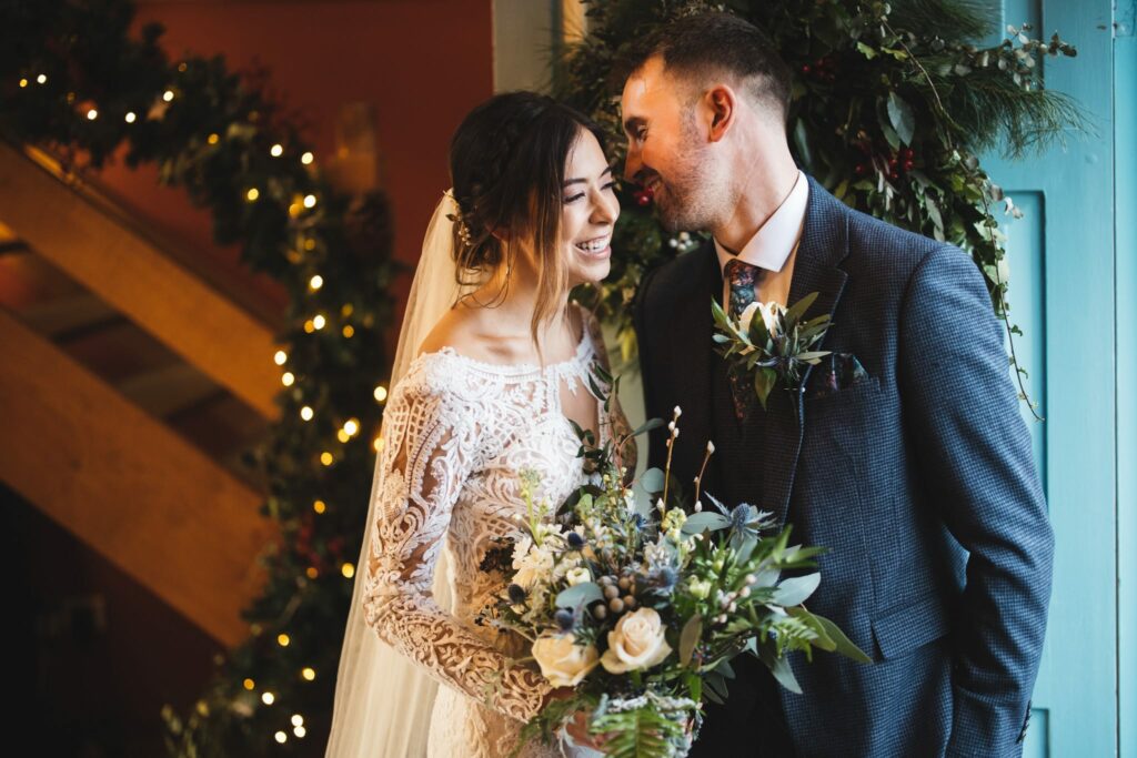 Winter wedding with christmas wreath and twinkly lights in background as bride and groom share a kiss on wedding day
