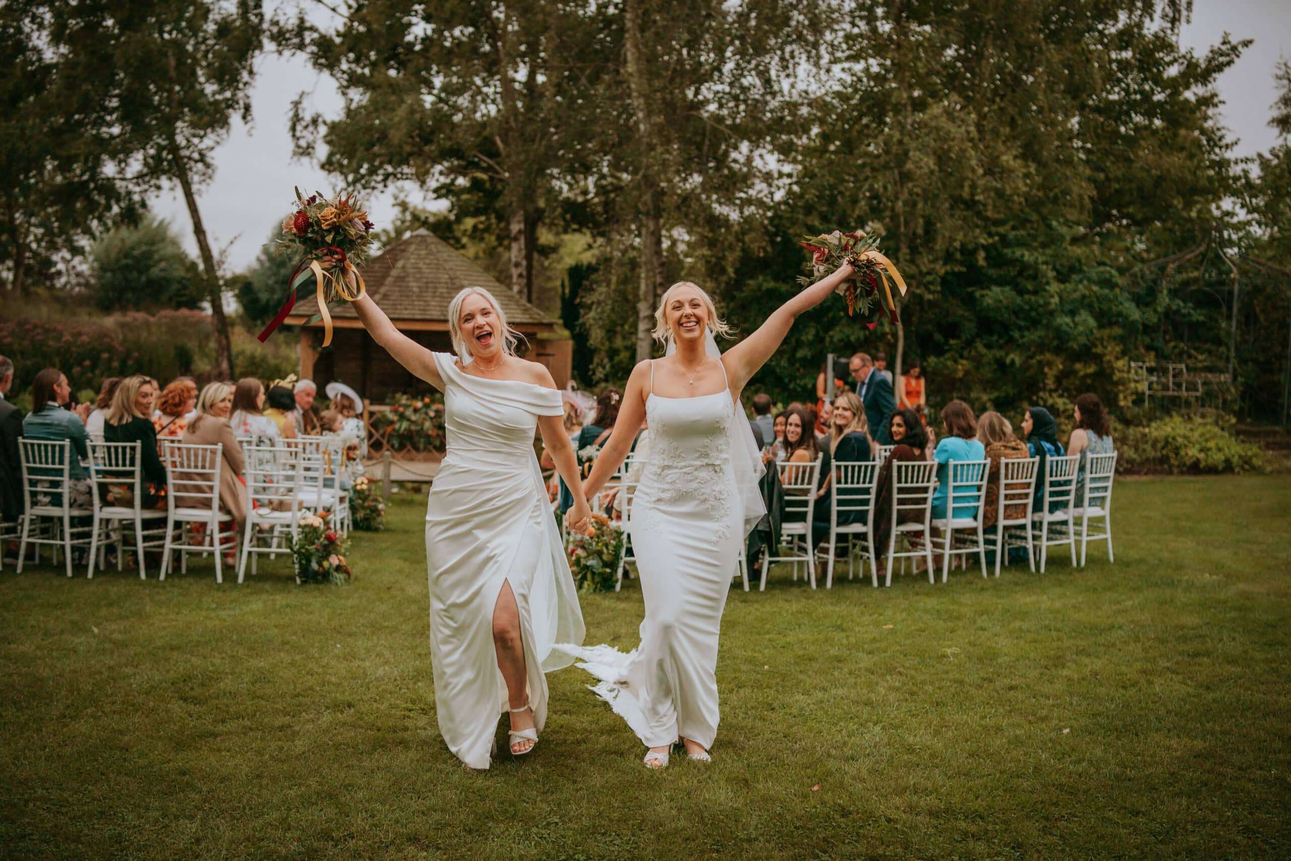Two brides in white wedding dresses leave garden ceremony raising their bouquets in the air as guests at the outdoor same sex wedding ceremony look on smiling