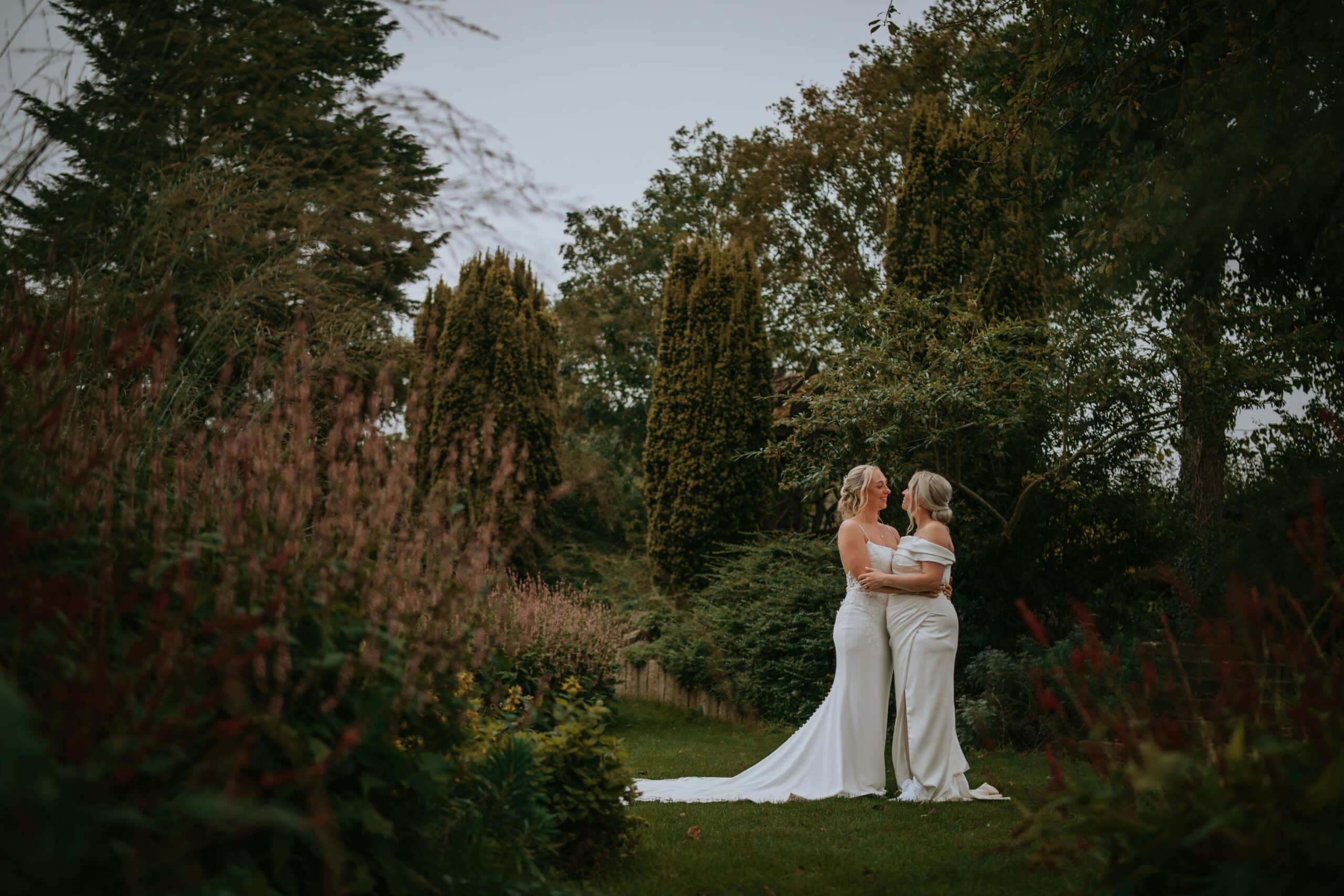 Two brides in beautiful white wedding dresses embrace in gardens of same at same sex wedding venue